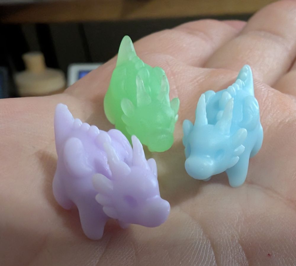 Three small dragon figurines in different colors, pastel purple, see through light green, and pastel blue, sitting on the palm of a hand