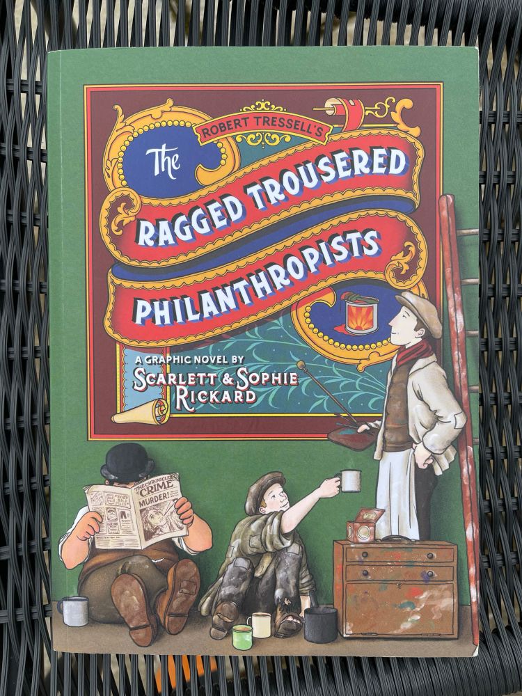 The front cover of The Ragged Trousered Philanthropists graphic novel. It is green and shows Owen standing back to look at a sign he’s painted (the title card of the book) while young Bert hands him a tin mug of tea. The foreman Crass is relaxing and reading a paper.