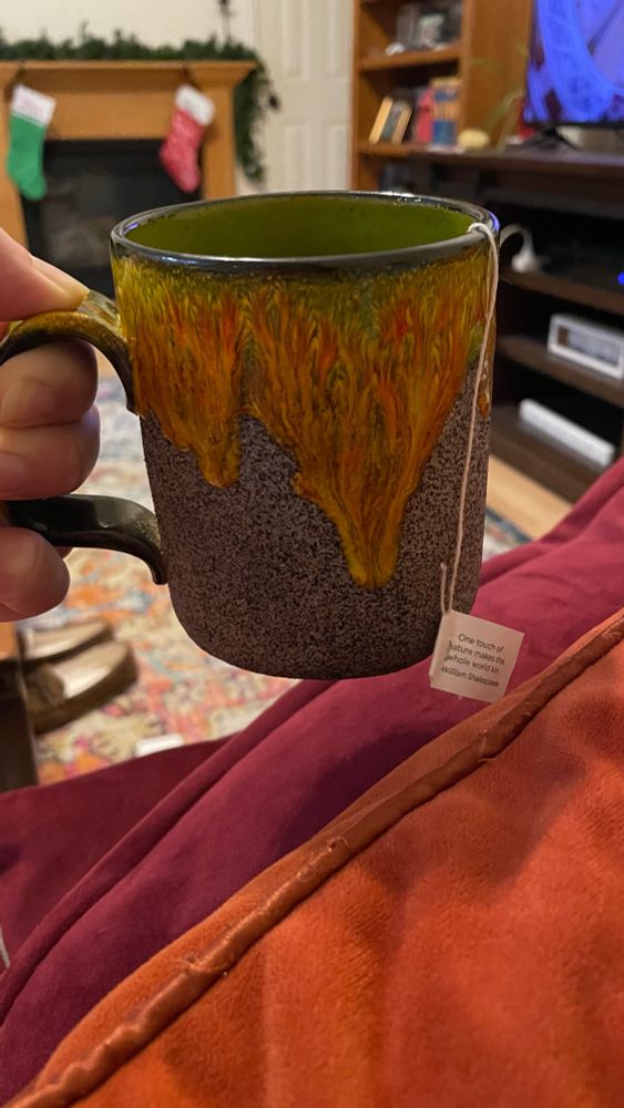 A mug of tea in a living room with a stack of pillows in the foreground. A fire place with stockings in the background. 