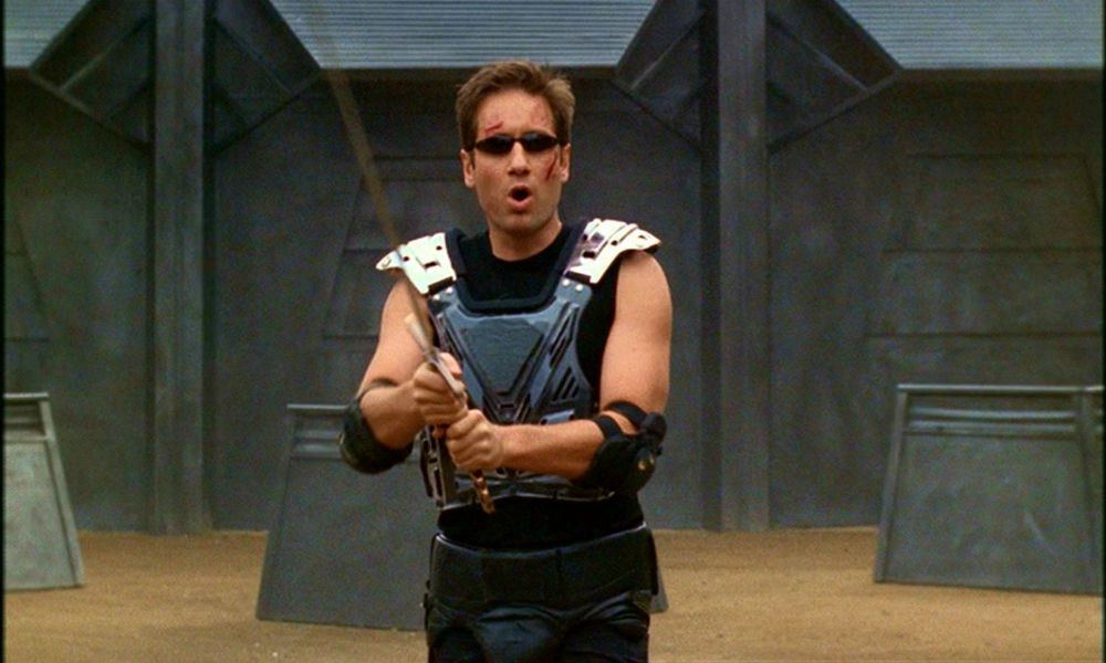 David Duchovny as Fox Mulder in The X-Files episode First Person Shooter. He has a black tank top on with metal armor vest on top. He has black elbow pads and is holding a sword. He also has slim black sunglasses on. 