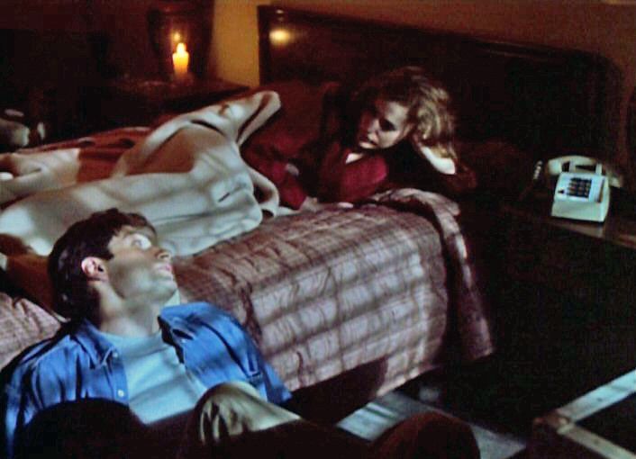 Mulder and Scully deep in conversation. Scully is laying in the motel bed in red pajamas with her hand propping her head up. Mulder is wearinf casual clothes and sitting on the floor with his back agains the side of the bed in the pilot episode of the The X-Files. 