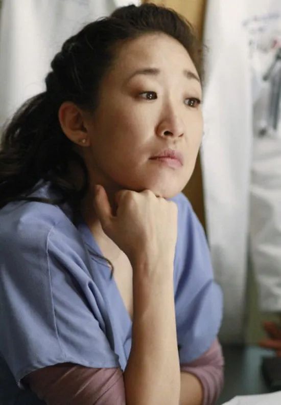 Cristina Yang from Grey’s Anatomy wearing light blue scrubs resting her chin on her fist. Hair in ponytail. 