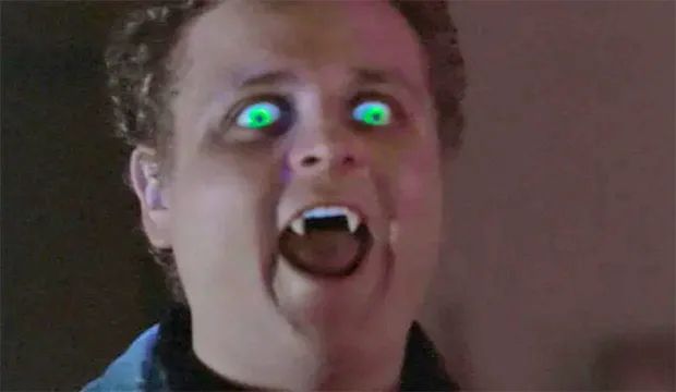 A teenage vampire with is mouth open. He has light brown curly hair, glowing green eyes, and fang teeth. 