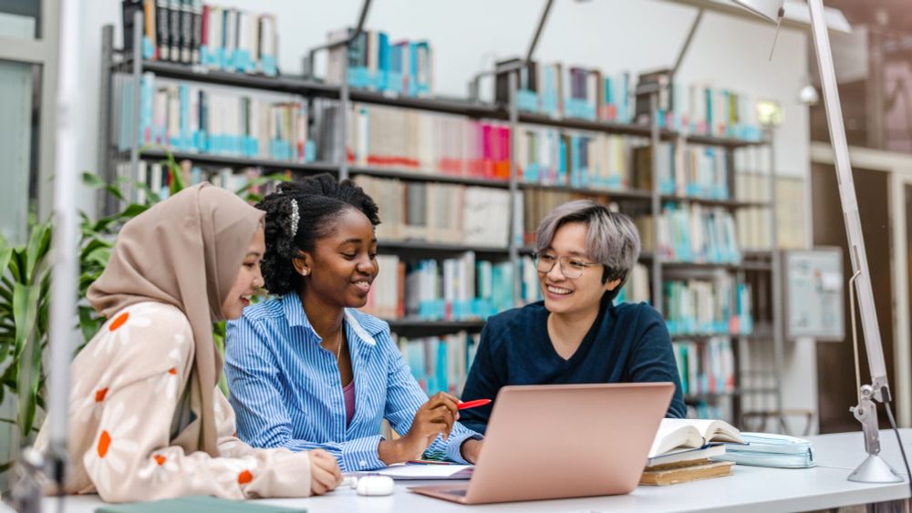 Three people sitting in a library, smiling, looking at a laptop.