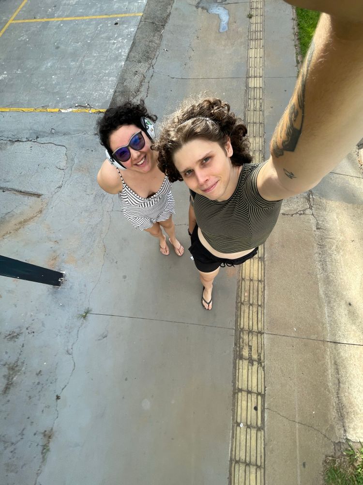 Duas pessoas na calçada. Uma está usando um macacãozinho listrado branco com preto e fones de cancelamento sonoro, a outra está usando um cropped verde com listras pretas e um shortinho preto. A câmera está em um ângulo de cima para baixo das duas 