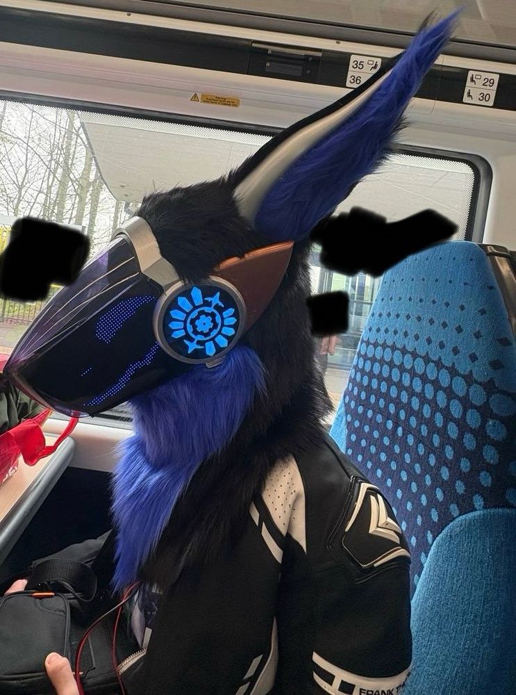 On a train, a blue and black furred protogen fursuit with blue leds and bronze spikes. 