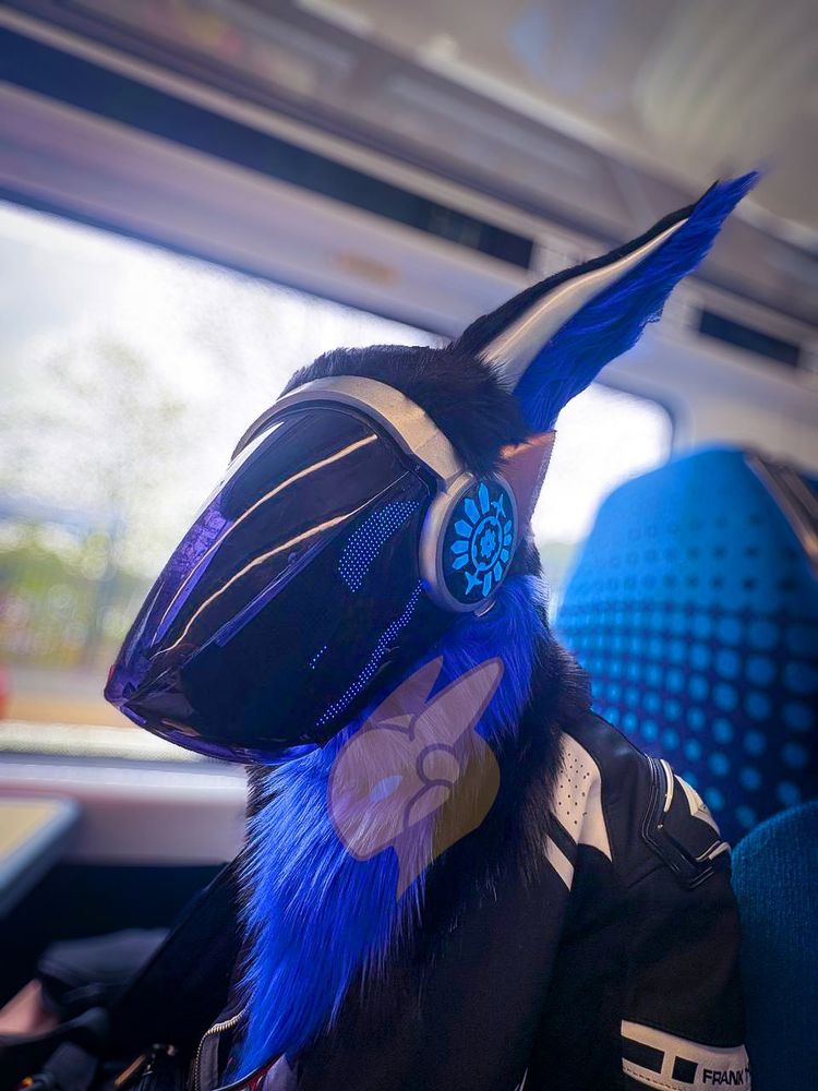 blue and black protogen fursuit with blue led face. Blue led patterned side panel and silver and bronze painted plastics.  Inside a modern train, camera angle is 3/4 side slightly from below.