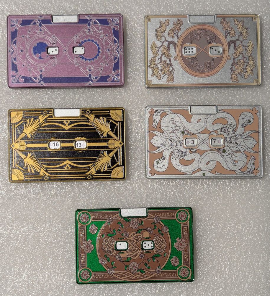 Five metal rectangular dice spinners, in various designs: purple arcane, black and gold art deco, green Celtic, and two silver dragons. The center has a pair of windows where the two numbers rolled are displayed.