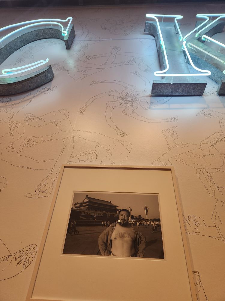 Photo of artist, Ai Wei Wei, with fuck painted on his chest. Photo is hanging in gallery with lighted neon C and K in frame.