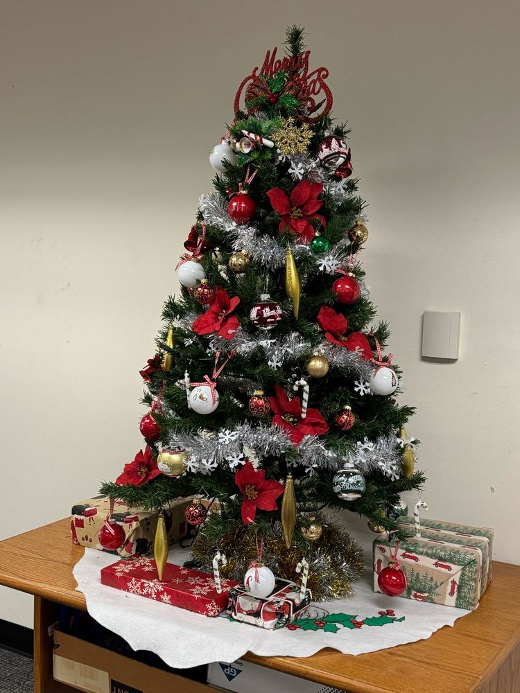 Christmas tree with reds poinsettias, white, and gold decorations surrounded by gift wrapped presents. The tree sits in a white tree skirt with holly leaves and berries.