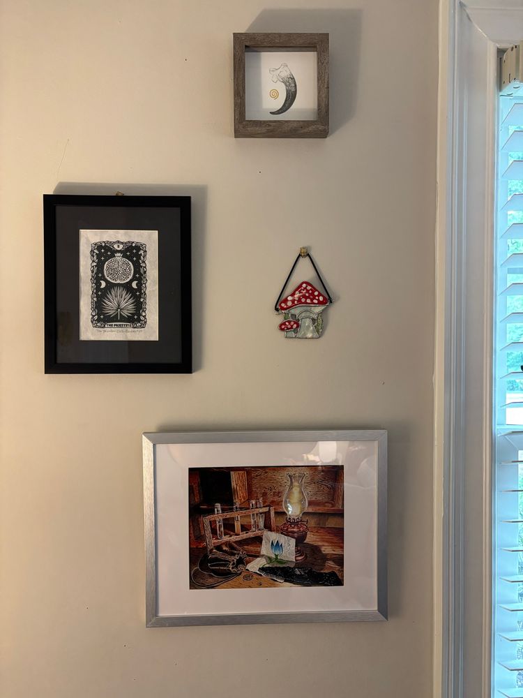 Photo of four art pieces hanging on a wall. 1. A graphite drawing of a talon, 2. A stained glass amanita mushroom, 3. A colored pencil still life depicting items from the Mistborn book series, and 4. A print of the high priestess tarot card 
