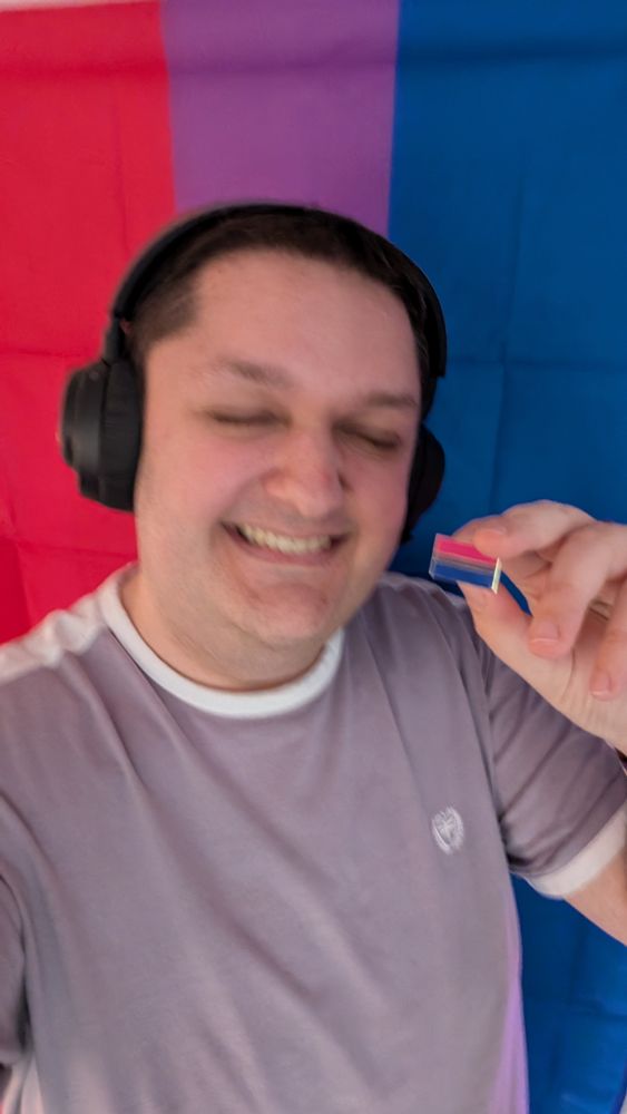 me, in front of a bisexual flag and holding a bisexual flag pin