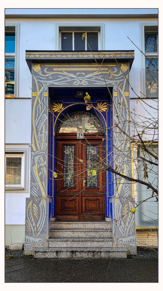 Das Bild zeigt den Eingangsbereich eines älteren Wohnhauses, frontal aufgenommen.
Im Mittelpunkt steht eine dunkle, zweiflügelige Holztür mit dekorativen Füllungen und farbigen Glaseinsätzen. Über der Tür befindet sich ein halbrundes Oberlicht, ebenfalls mit ornamentiertem Glas. Der Türrahmen und das Portal sind reich gestaltet: grau-blaue und goldene Mosaik- und Reliefornamente mit floralen und geschwungenen Motiven umgeben den Eingang und verleihen ihm einen jugendstilartigen Charakter.
Mehrere Steinstufen führen zur Tür hinauf. 