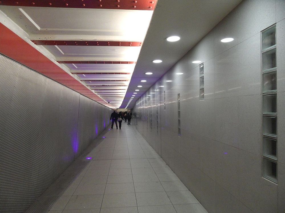 Langer, mit Neonlicht beleuchteter Tunnel. Hinten der Ferne kann Passanten sehen.
