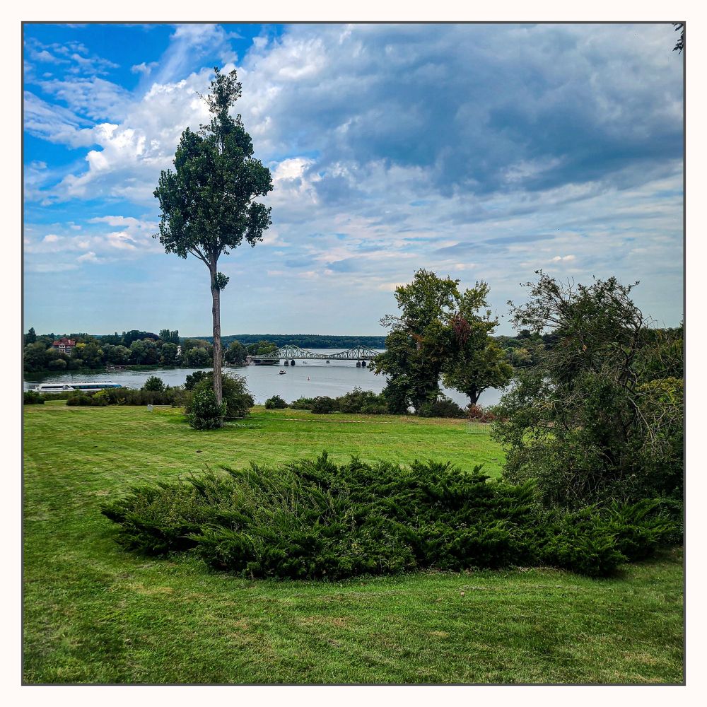 Wolken am blauen Himmel links ein Bäumchen, dahinter ein Gewässer und in der Ferne eine Brücke und Uferlinien.