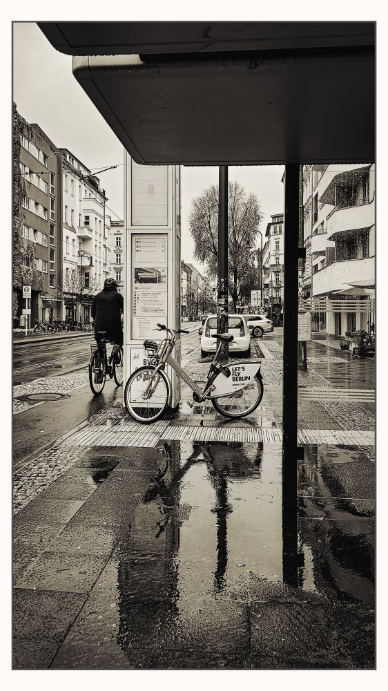 Wartehäuschen an einer Straßenbahnhaltestelle. Auf dem Plattenboden hat sich eine Pfütze gebildet, in dem die Kreise der Regentropfen zu sehen sind. An der Fahrplansäule lehnt ein Fahrrad. Links daneben radelt gerade ein Radfahrer vorbei. 
Das Foto ist monochrom. 