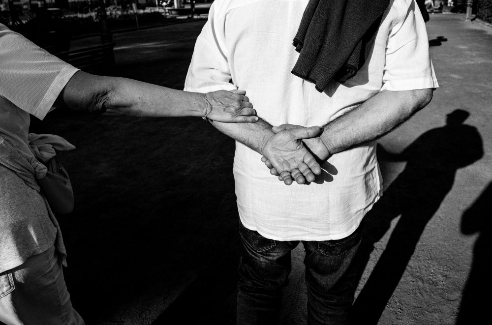 A man's hands clasped behind his back, with a woman holding on to them. Their shadows stretch ahead of them. Black & white. 