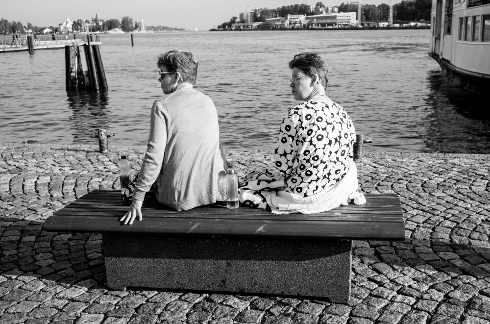 A black and white photo of two women on a bench, turning at the same angle, with the water in the background.