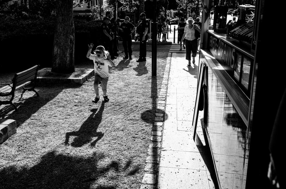 A black and white picture of a path, with a small child running in the direction of the camera, lit from behind, with their shadow showing their raised hands. 