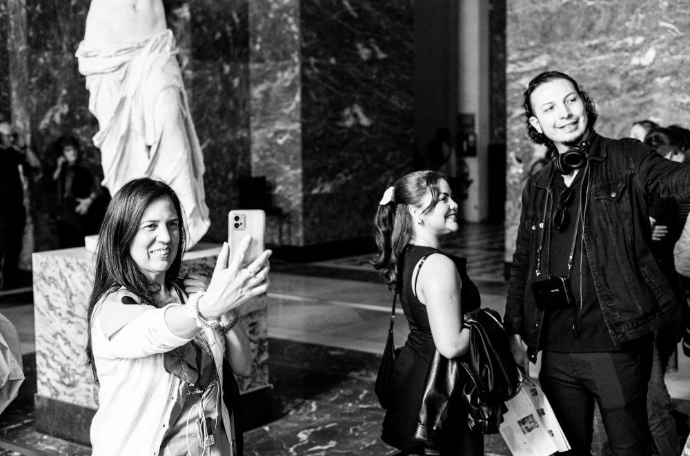 A black and white picture of several people posing taking selfies in front of the famous Venus de Milo sculpture in the Louvre, Paris, France.