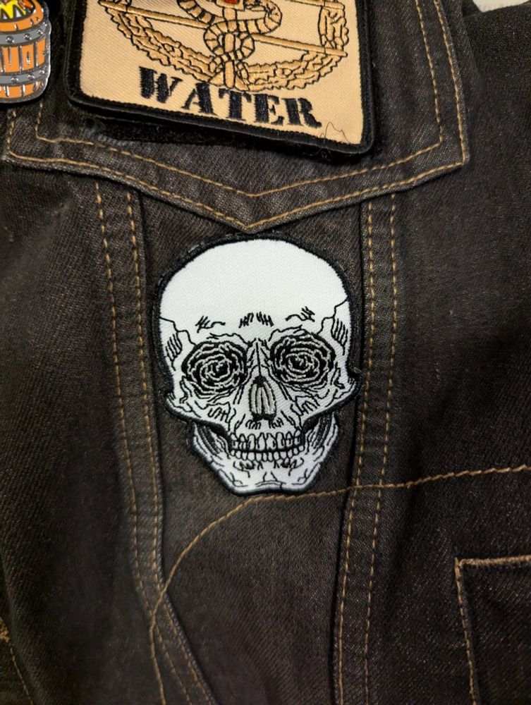 Tired looking skull patch with glow in the dark rose eyes on a denim vest under another patch with the combat medic symbol & the words "Motrin" and "Water".