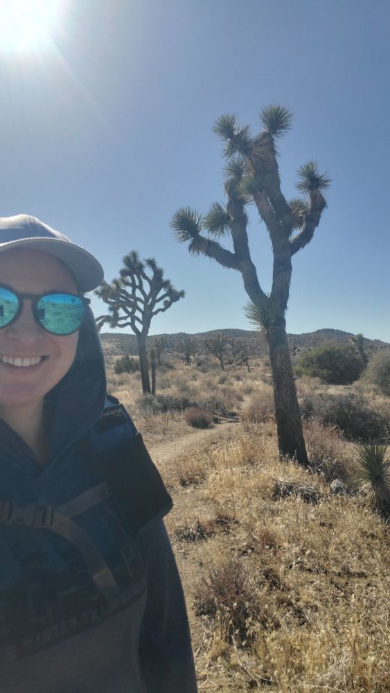 A selfie of Katie with two Joshua Trees in the background. The sky is bright blue, the ground is scrubby, and two large Joshua Trees dominate the field of view. Katie is wearing bright blue sunglasses and is facing away from the sun