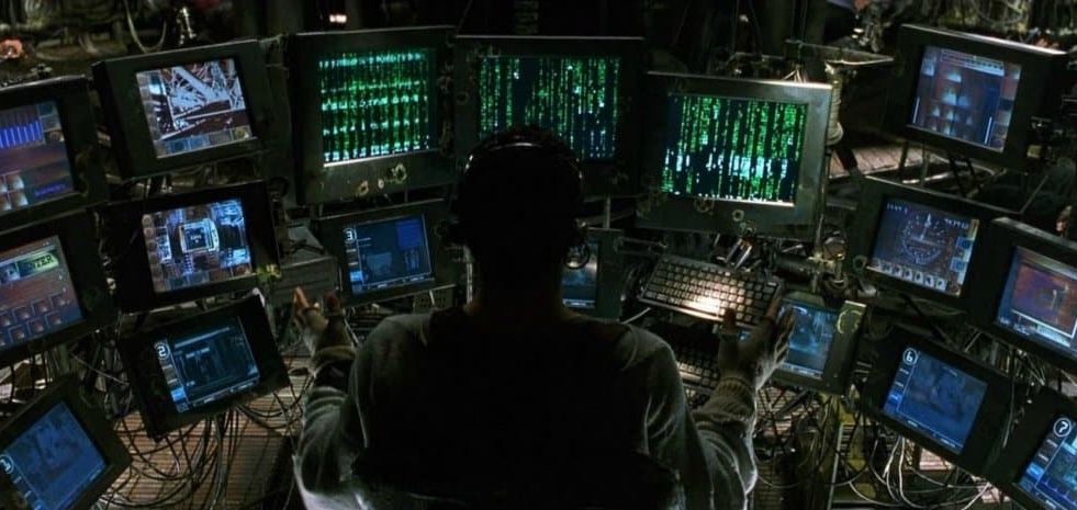 The character "Tank" from the movie "The Matrix" sitting in front of like, twelve monitors, all showing different tech stuff.