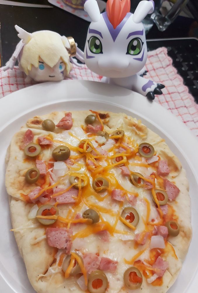 A photo of a pizza made with garlic naan bread, on a white plate. Behind the plate are 2 digimon, a Gomamon figure and a small Lucemon plushie.