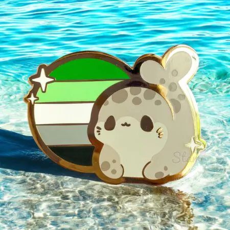 A photo of an enamel pin with a circular aromantic flag design with a chibi art style grey spotted seal beside it. 
