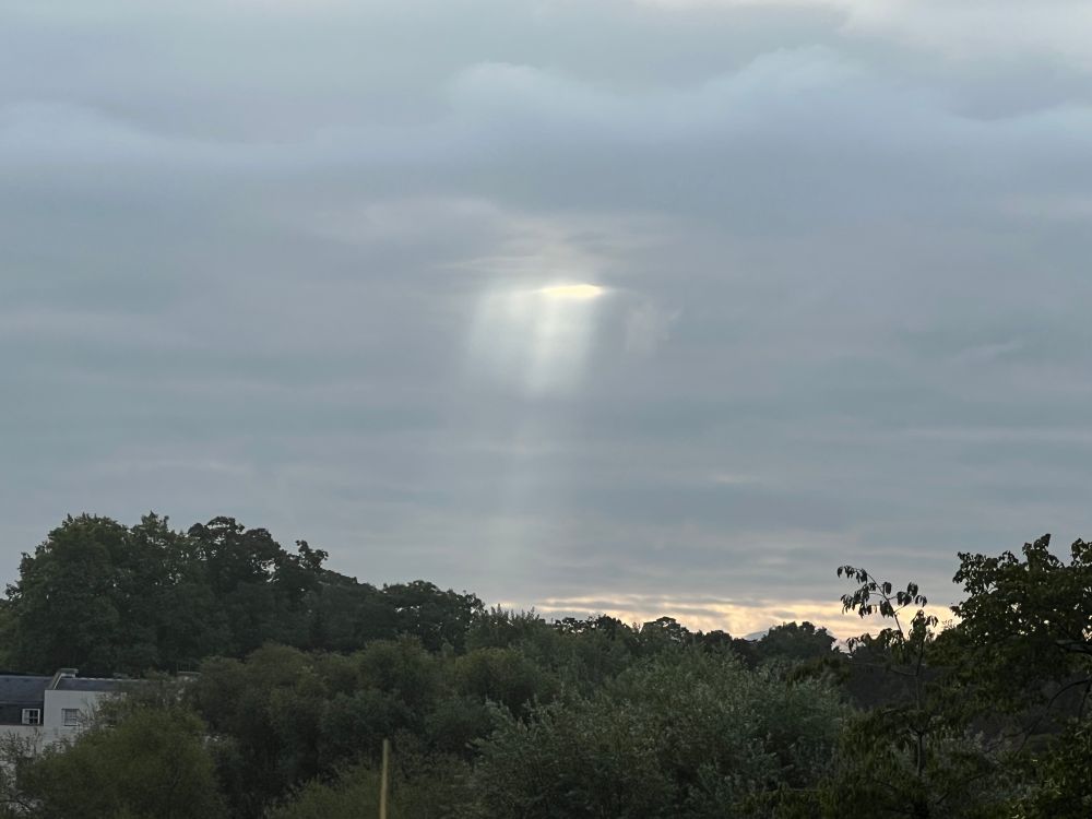 A photo of an overcast day. The sky has thick grey clouds. There is a small hole in the clouds through which a thin shaft of sunlight shines. 