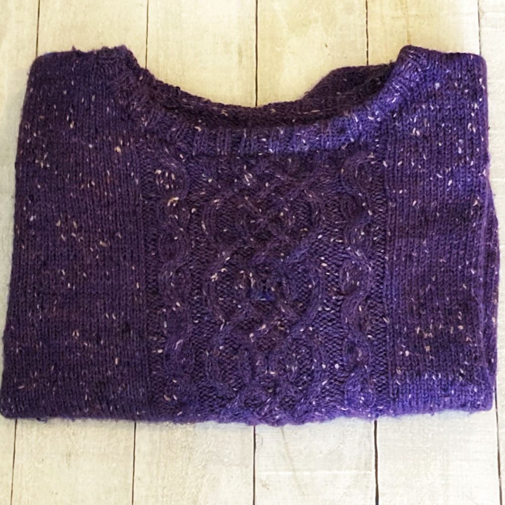 Folded handknitted jumper in purple tweed with a cable pattern on a white wooden background.