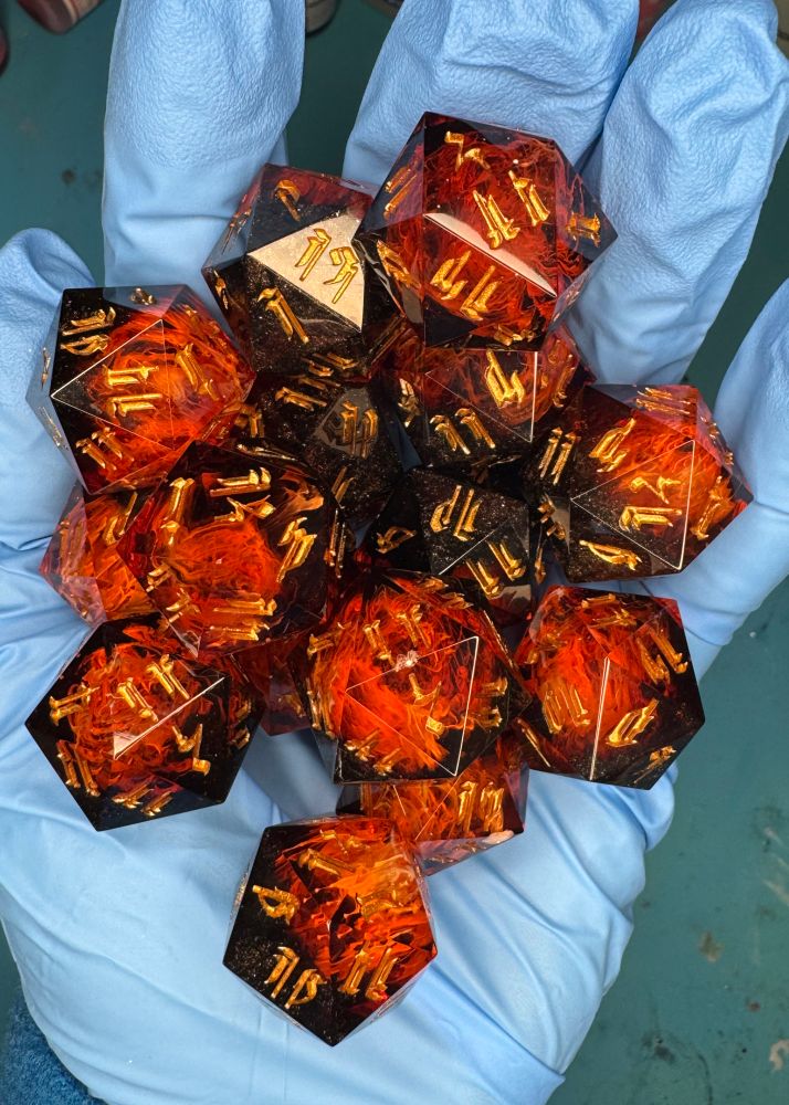 A handful of 15 individual d20 dice that look like fire and have gold numbers in the Infernal alphabet. The hand is wearing a blue nitrile glove. 