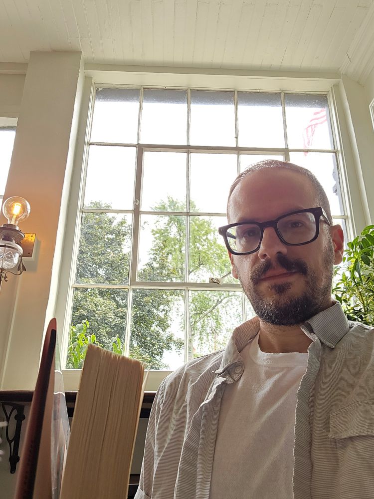 Me sitting in a café with a tall windoe behind me. The light is soft and white with a hint of yellow from a wall sconce behind me. I'm wearing a white t-shirt, glasses, and holding a book.