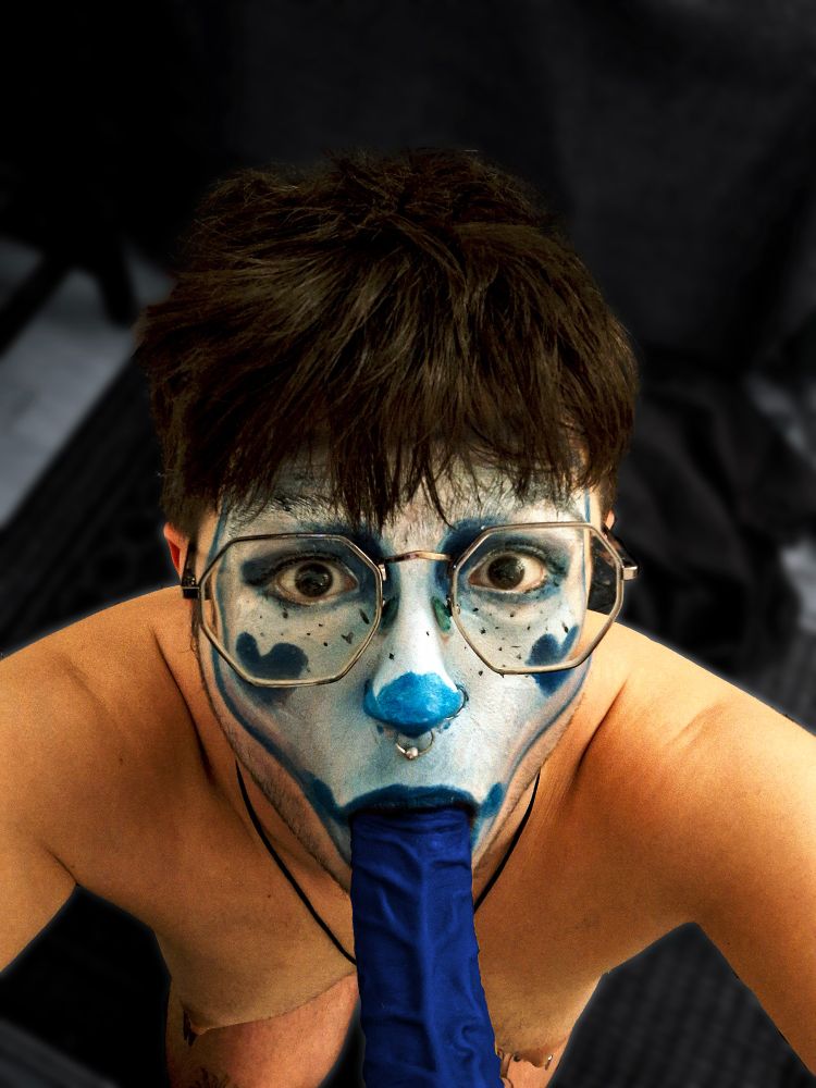 A clown... girl? looks into camera with their mouth around a blue veiny dildo. Their face is painted with a mask of whiteface clown makeup, with blue heart details and freckles. 
