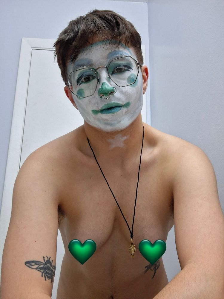 A clown in full whiteface with blue-teal monochromatic features looks down at the camera, bemused. There is a white star painted on their throat in clown makeup, and their nipples are censored by green emoji hearts. 
The background is a light blue wall with a white door.