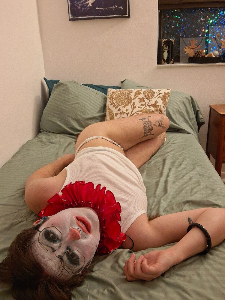 A clown looks up at you from a mattress. Background is a bedroom - a windowsill with botanical objects on it and a wooden side table