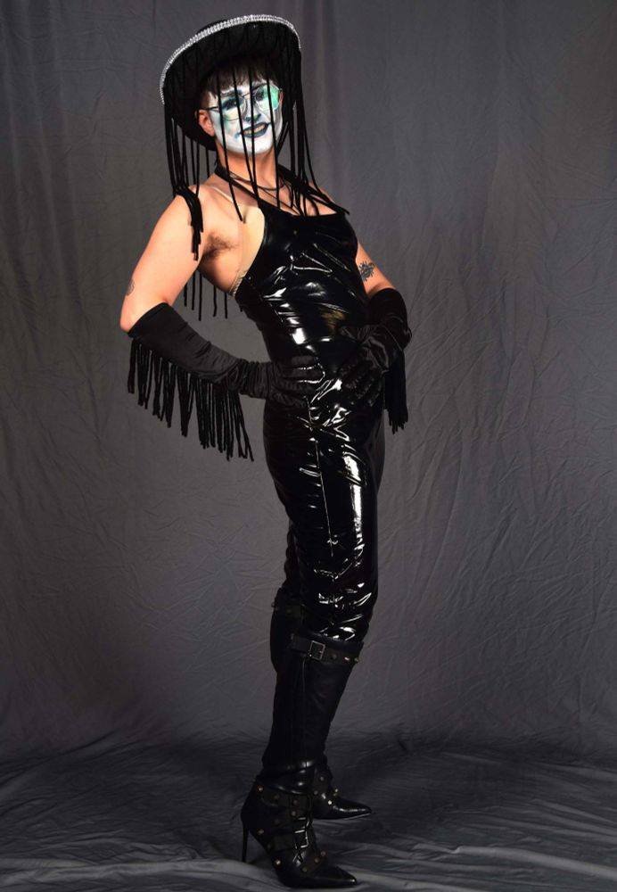 An androgynous clown in a full pvc bodysuit, fringe rimmed cowboy hat, fringed opera gloves and unfringed leather boots, all black. They lean back while standing and smile at camera. 
Background is a gray photoshoot tarp, slightly winked. 