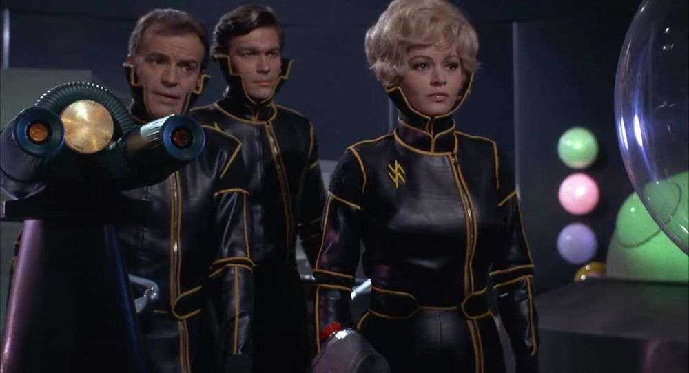 Scene from "Planet of the Vampires" (1965) directed by Mario Brava. Three people in severe black leather scifi military uniforms with gold trim and very high upraised collars stand before a mounted ray-gun. Left to right: Captain Mark Markacy (Barry Sullivan), Sgt. Harry Gash (Federico Boido), and Lt. Tiona (Evi Mirandi).  