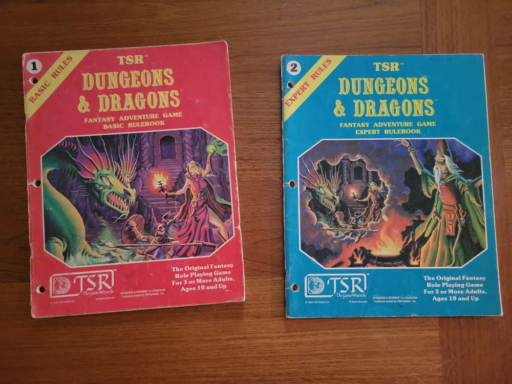 Dungeons & Dragons Basic Rules and Expert Rules softcover books, 1981 editions. Basic Rules has a red cover with an illustration of a sorceress and a knight with a spear fighting a green dragon. Expert Rules has a blue cover and shows a wizard casting a spell over a cauldron. Smoke rises from the cauldron forming a frame, in which is the illustration from the Basic Rules book.