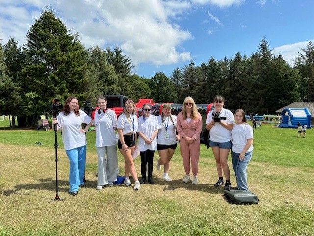 Seven young people wearing white T‑shirts and casual pants or shorts—some holding camera gear—standing in a line on a sunlit grassy field alongside Commissioner Nicola Killean in a pink suit and sunglasses; behind them are trees, inflatable play structures, and a partly cloudy blue sky.