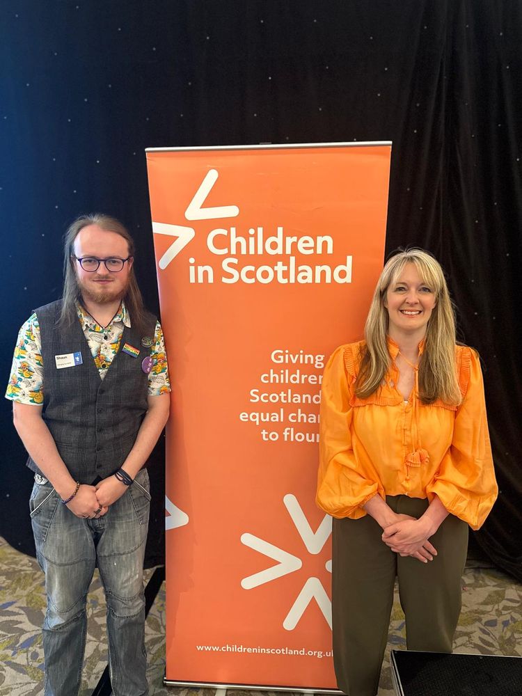 Wearing an orange shirt and khaki trousers, Nicola Killean, the Children and Young People's Commissioner is standing with a young person called Shaun, who is wearing a waistcoat and jeans. Between them is a sign advertising the organisation called Children in Scotland. 