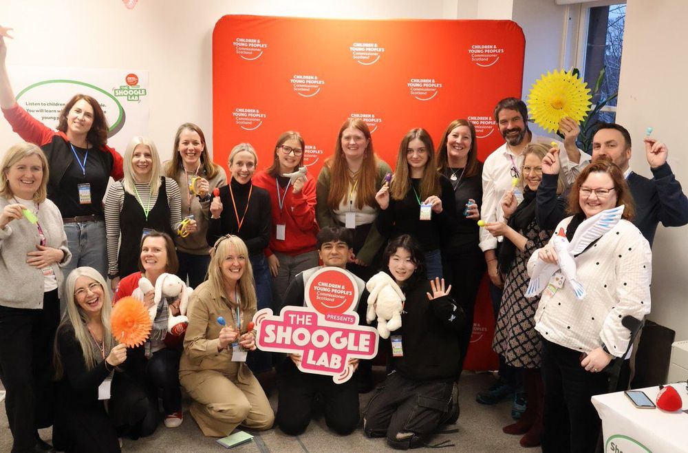 A large group of Young Advisors, the Commissioner’s team, and education leaders pose together at the end of the Shoogle Lab event. Everyone is smiling and holding up shoogle props like maracas, soft toys, and colourful objects. Two young people kneel at the front holding a “The Shoogle Lab” sign. Behind the group is a red Children and Young People’s Commissioner Scotland backdrop.