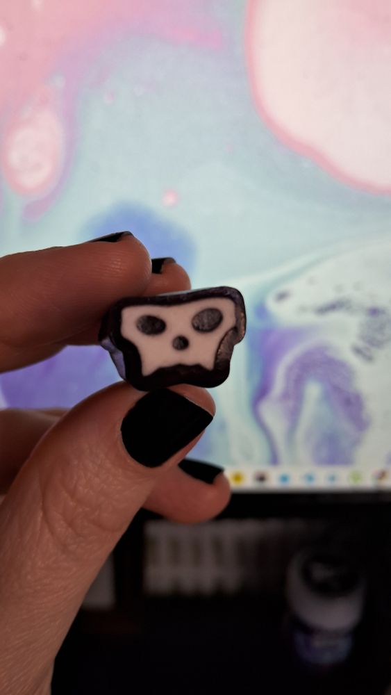 foto da minha mão segurando uma bala sabor amora com capim-limão que é uma caveira estilizada em preto e branco. minhas unhas estão curtas e pintadas de preto. ao fundo aparece meu monitor desfocado com o desktop e um papel de parede com formas abstratas nas cores azul, rosa e roxo.