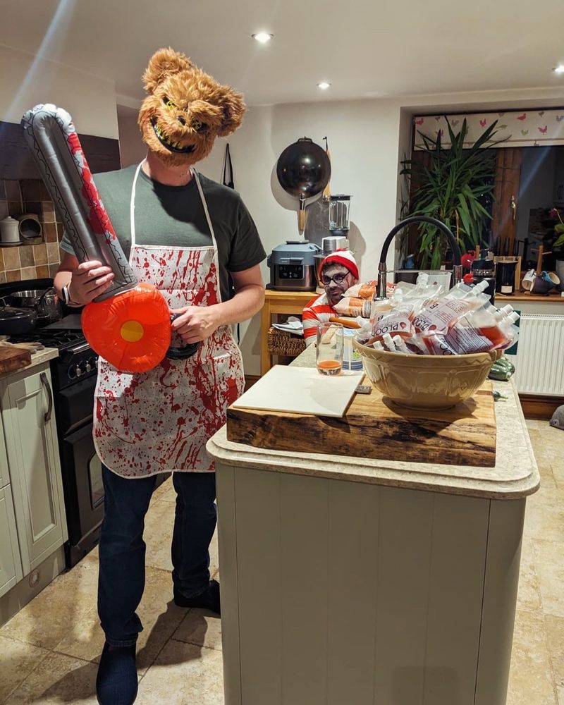 A person in a kitchen dressed as a killer bear with a blow up chainsaw. Next to a large bowl of fake blood bags filled with cocktails. With a Wally hiding in the background. 