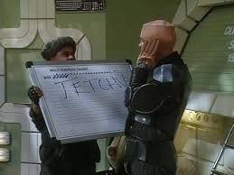 Lister from Red Dwarf holding up a whiteboard so that Kryton can see he’s being called “TETCHY” despite blocking his ears. 