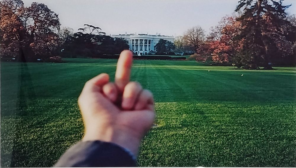 Ein Foto vom weißen Haus. Davor eine Hand, welche dem weißen Haus den Mittelfinger zeigt.