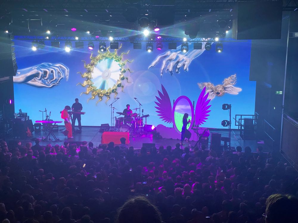 Magdalena bay concert stage Mika standing jn front of a oval screen flanked by white angel wings
