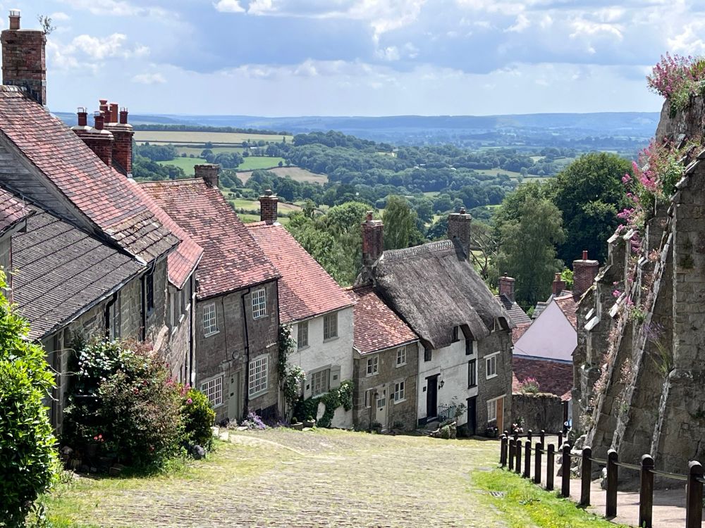 Gold Hill in Shaftesbury, sloping cobbles street with lots of cottages and green hills and blue sky in the background 