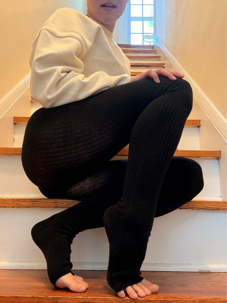 mal perches on a flight of stairs wearing a cropped white sweatshirt and black tights that leave their toes exposed. they are on tiptoe, mouth slightly open.