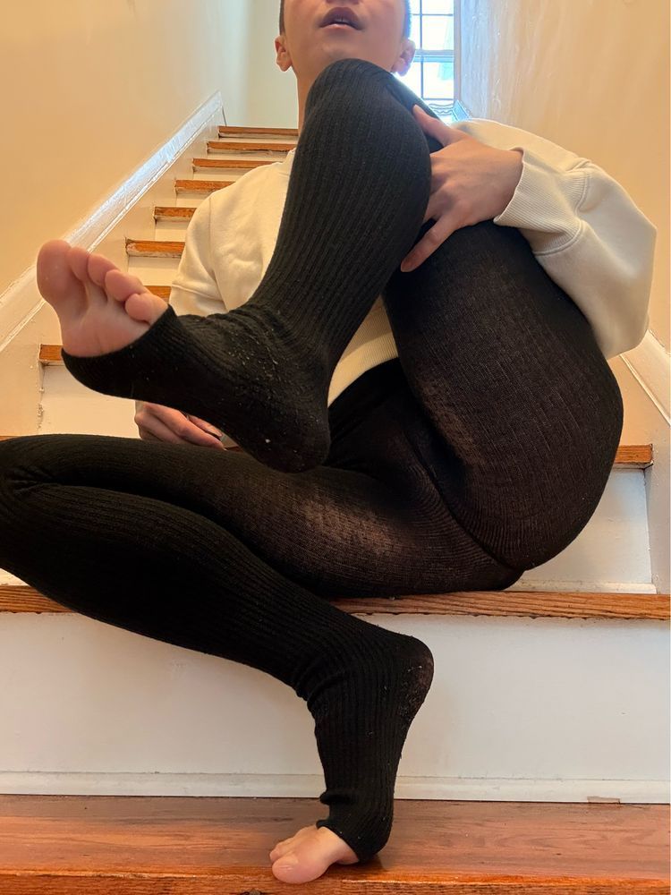 mal perches on a flight of stairs wearing a cropped white sweatshirt and black tights that leave their toes exposed. one knee is drawn up to show that they aren't wearing anything under their tights.